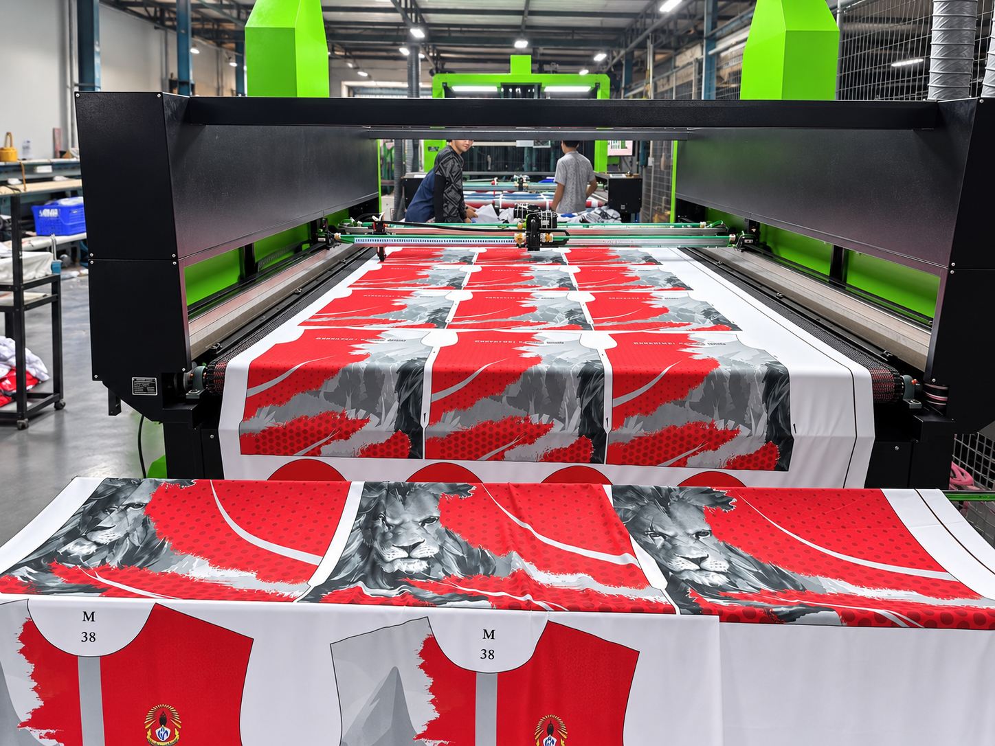 A large flatbed gantry machine with light bars and an operator screen, printed fabric on the bed and two operators standing in front